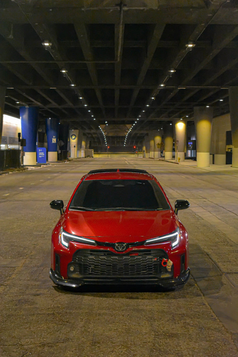 Load image into Gallery viewer, Glossy black V2 front lip installed on a red Toyota GR Corolla (E210, 2023-2024) parked under a bridge.