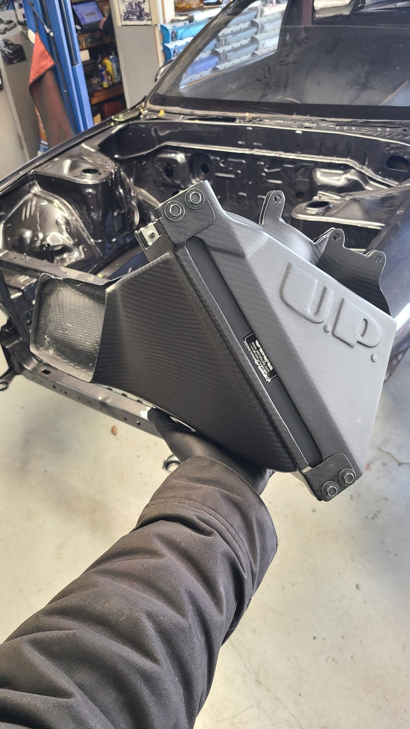 Load image into Gallery viewer, U.P.G R32 GTR oil cooler with black carbon fiber ducting held in hand, front of car chassis in background.