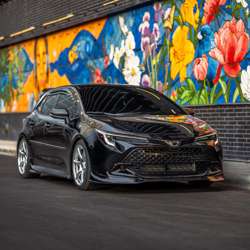 Load image into Gallery viewer, Gloss black front lip on a black 2023+ Toyota Corolla Hatchback (E210 Kouki) parked by a colorful mural.
