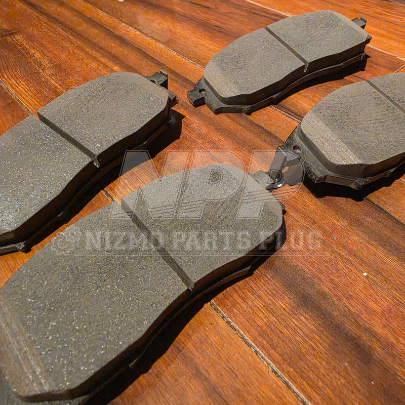 Load image into Gallery viewer, Four grey front brake pads for Nissan Stagea RS4-V (11/98) on a wood background. Good condition with visible wear.