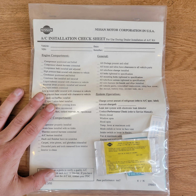 Load image into Gallery viewer, New old stock S13 240SX A/C installation check sheet and oil packet in a sealed plastic bag held by a hand.