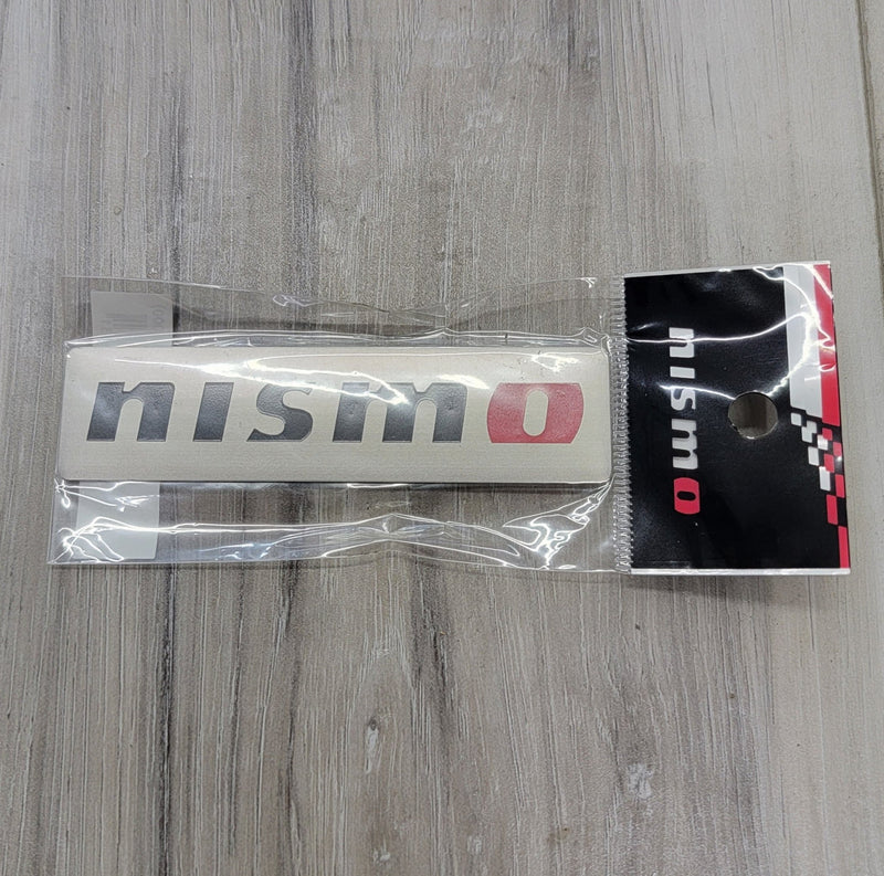 Load image into Gallery viewer, New brushed aluminum Nismo emblem badge in original packaging. Fits various Nissan models including R32, R33, R34.
