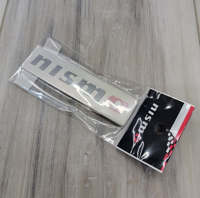 Load image into Gallery viewer, New Nismo brushed aluminum emblem badge in clear retail packaging, suitable for various Nissan models.