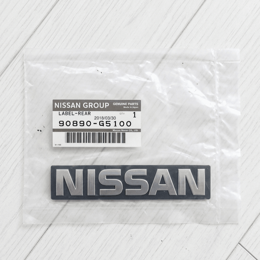 New genuine Nissan 1200 rear emblem in silver and black, sealed in original OEM packaging with part label.