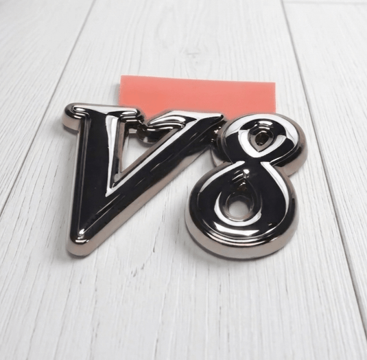Chrome V8 emblem for Nissan FGY33 Cima. Shiny metallic finish on a white wood surface.