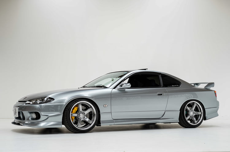 A silver Nissan Silvia S15 coupe with custom multi-spoke wheels, a lowered suspension, and a rear spoiler, photographed in a side profile against a white studio background.