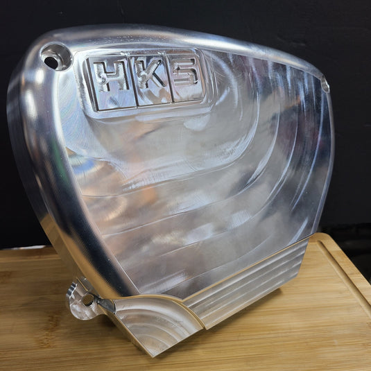 Polished billet aluminum HKS front timing cover for RB26 engines, featuring CNC-machined texture.