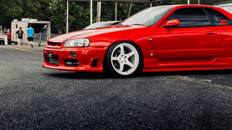 A bright red custom Nissan Skyline R34 coupe with white multi-spoke wheels, parked on asphalt at an outdoor car show. A dark brown canopy tent with 'none like mine' branding is visible in the background, along with trees and a cloudy sky.